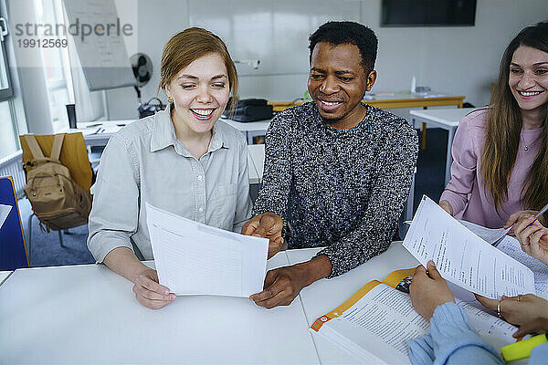 Lächelnde multiethnische Studenten mit Notizen  die gemeinsam am Schreibtisch im Klassenzimmer lernen