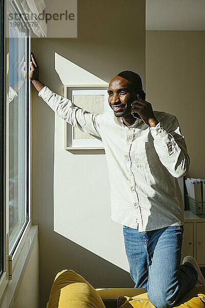 Lächelnder Mann  der zu Hause im Sonnenlicht am Fenster steht und mit dem Handy telefoniert