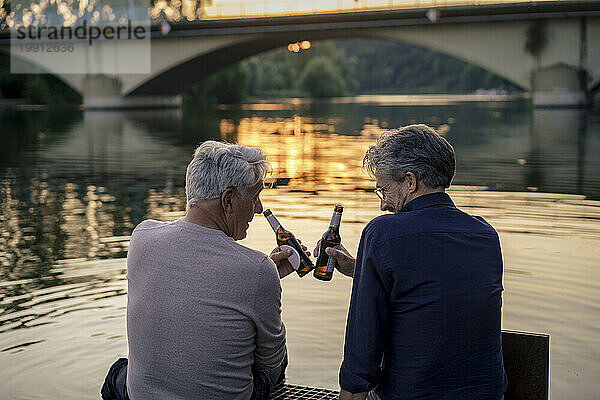 Ältere Freunde toasten bei Sonnenuntergang Bierflaschen am Wasser