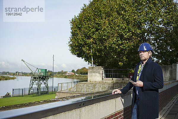 Reifer Architekt steht mit Smartphone auf Brücke in der Nähe des Flusses