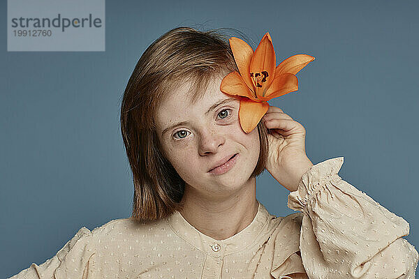 Mädchen mit Behinderung hält orangefarbene Orchidee hinter dem Ohr vor blauem Hintergrund