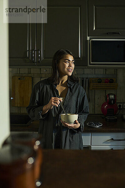 Lächelnde Frau hält eine Schüssel Frühstück in der Hand und steht zu Hause in der Küche