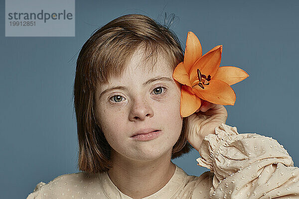 Mädchen mit Down-Syndrom hält orangefarbene Orchidee hinter dem Ohr vor blauem Hintergrund
