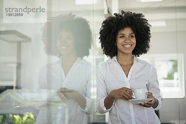 Smiling businesswoman holding tea cup leaning on glass wall at office