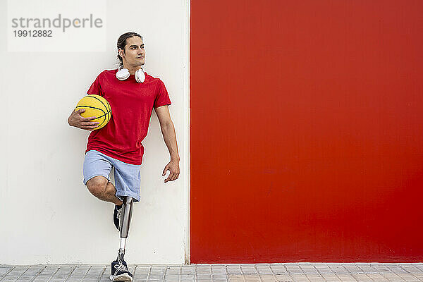 Lächelnder Mann mit Behinderung hält gelben Basketball in der Hand und lehnt an der Wand