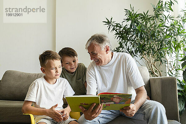 Lächelnder älterer Mann liest zu Hause im Wohnzimmer ein Buch für seine Enkel