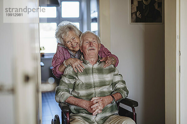Glückliche Seniorin umarmt Mann  der zu Hause im Rollstuhl sitzt
