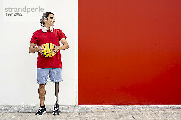 Lächelnder junger Mann steht mit Beinprothese und hält gelben Basketball vor der Wand