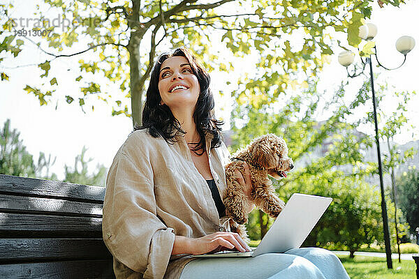 Lächelnder Freiberufler hält Hund in der Hand und sitzt mit Laptop auf Parkbank