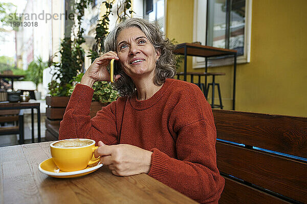 Glückliche Frau  die im Straßencafé mit dem Smartphone spricht und eine Kaffeetasse in der Hand hält