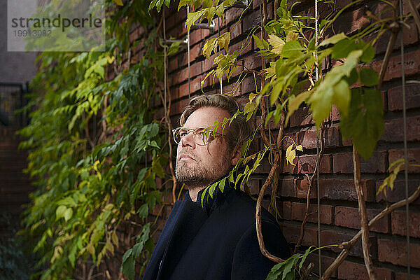 Contemplative mature businessman leaning on brick wall overgrown with plants