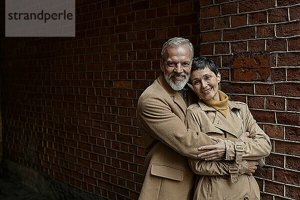 Happy couple standing against brick wall  cuddling each other and looking at camera