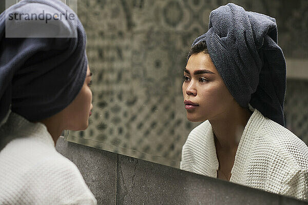 Junge Frau trägt ein Handtuch auf dem Kopf und schaut zu Hause in den Spiegel