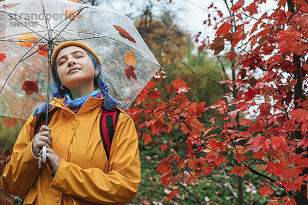 Lächelnde Frau hält Regenschirm neben Baum im Herbstpark