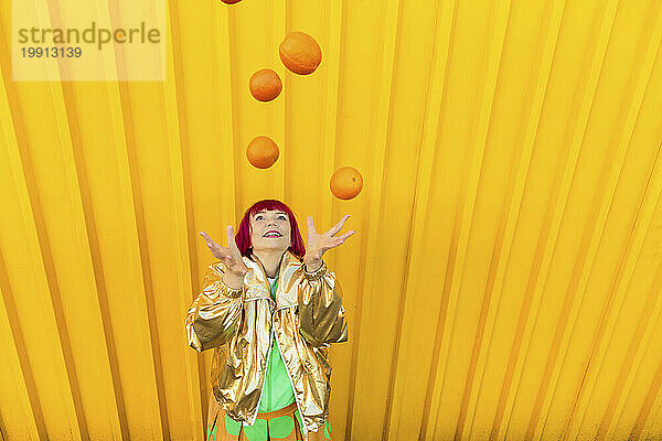 Glückliche Seniorin jongliert mit Orangen vor gelber Wand