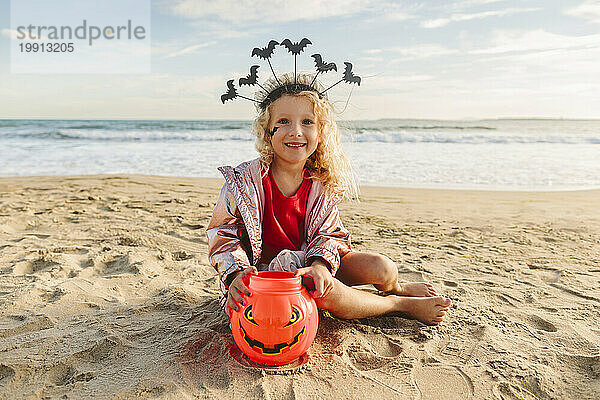 Lächelndes Mädchen sitzt mit Halloween-Kürbisspielzeug auf Sand am Strand