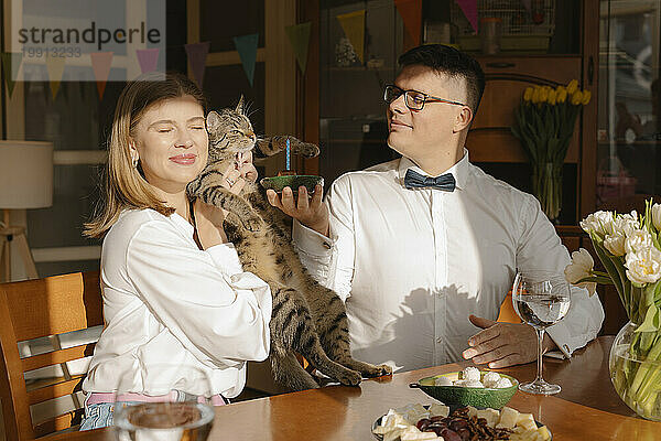 Young woman embracing cat with man holding birthday cake at home