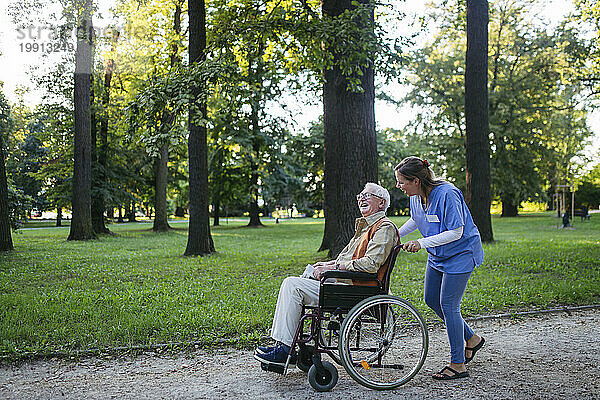 Glücklicher Mitarbeiter im Gesundheitswesen  der mit einem älteren Mann spaziert  der im Rollstuhl in der Nähe von Bäumen im Park sitzt