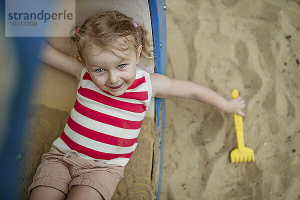 Glückliches kleines Mädchen mit Spielzeugrechen  das im Klettergerüst auf dem Spielplatz liegt