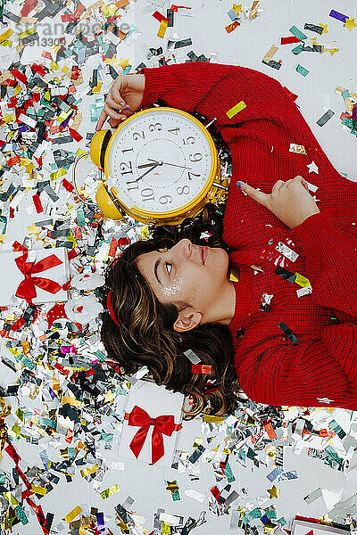 Smiling girl holding clock and lying on confetti