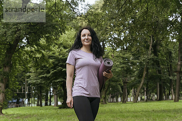 Lächelnde Frau hält Trainingsmatte in der Hand und steht im Park