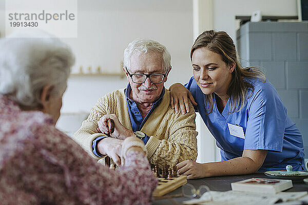 Lächelnder Mitarbeiter des Gesundheitswesens  der zu Hause Schach mit einem älteren Paar spielt
