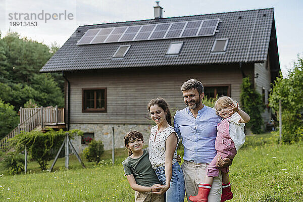 Glückliche Familie steht vor ihrem Einfamilienhaus mit Sonnenkollektoren auf dem Dach