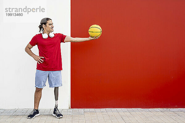 Glücklicher Mann mit Beinprothese hält gelben Basketball an der Wand