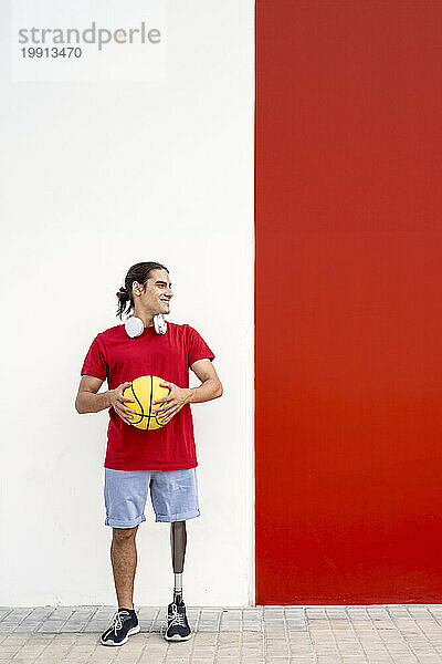 Glücklicher junger Mann mit Beinprothese hält gelben Basketball vor der Wand