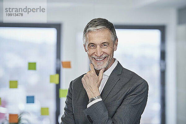 Happy senior businessman with hand on chin standing in office