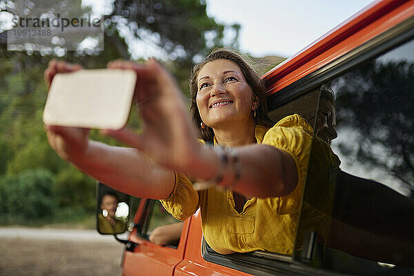 Lächelnde Frau macht ein Selfie mit ihrem Smartphone und lehnt sich aus dem Fenster ihres Wohnmobils