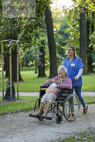 Glückliche Mitarbeiterin im Gesundheitswesen geht mit einer älteren Frau im Rollstuhl im Park spazieren