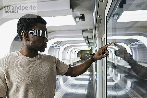 Junger Mann mit Cyberbrille berührt Glasscheibe mit dem Zeigefinger