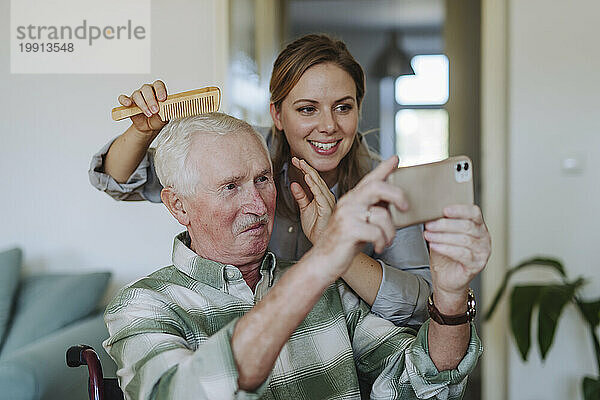 Fröhlicher Mitarbeiter im Gesundheitswesen kämmt die Haare eines älteren Mannes  der zu Hause ein Selfie macht