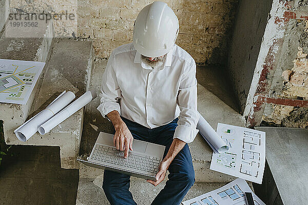 Architect using laptop with blueprints on staircase at site
