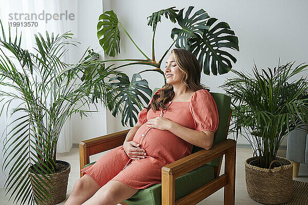 Smiling pregnant woman with hands on stomach sitting on armchair at home