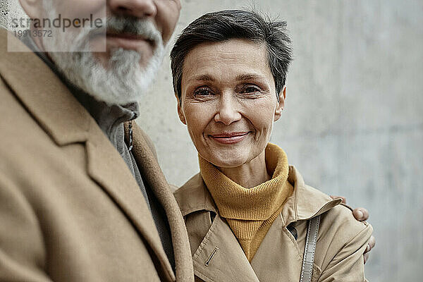 Erwachsene Frau blickt in die Kamera und steht neben ihrem Mann mit grauem Bart im Freien