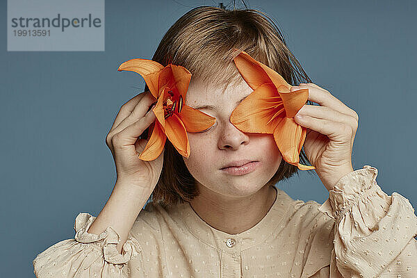 Teenagermädchen bedeckt ihre Augen mit orangefarbenen Orchideenblüten vor blauem Hintergrund