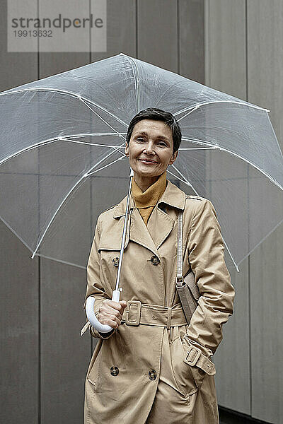 Charmante Frau im beigen Regenmantel steht unter einem Regenschirm und blickt in die Kamera