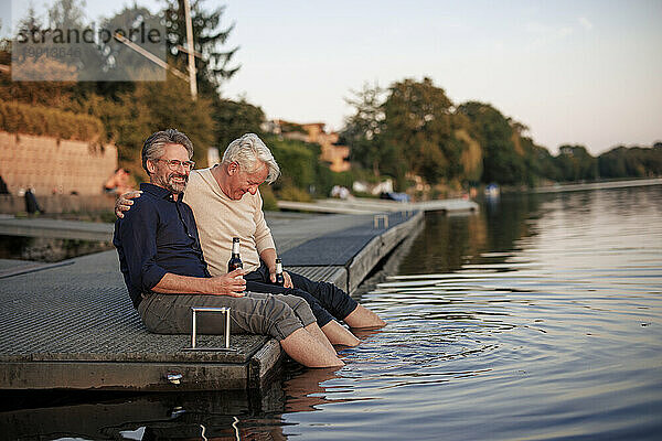 Glückliche ältere Freunde sitzen mit Bierflaschen auf einem Steg in der Nähe des Sees