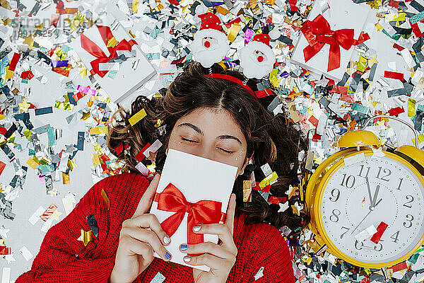 Mädchen versteckt Gesicht mit Geschenkbox und liegt auf Konfetti neben der Uhr