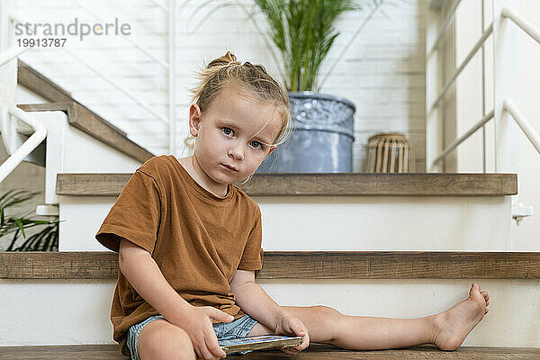 Unschuldiger blonder Junge mit Handy sitzt zu Hause auf der Treppe