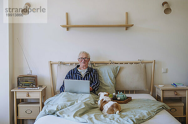 Lächelnder älterer Mann sitzt mit Laptop und Hund zu Hause im Bett