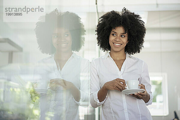 Smiling businesswoman with tea cup leaning on glass wall at office