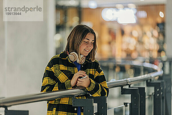 Lächelndes Teenager-Mädchen mit kabellosen Kopfhörern steht in der Nähe des Geländers im Einkaufszentrum