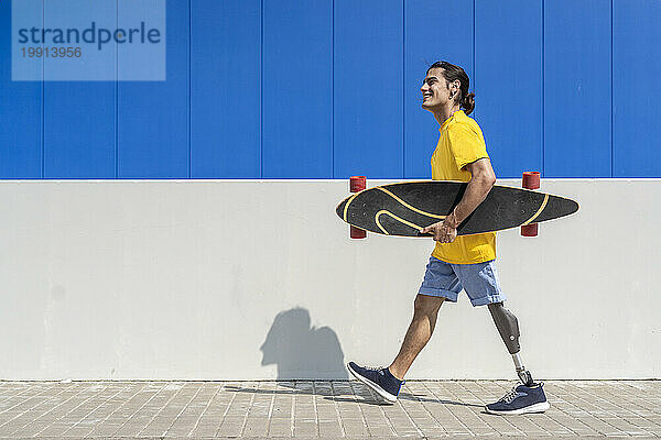 Glücklicher junger Mann mit Skateboard und Beinprothese vor der Wand