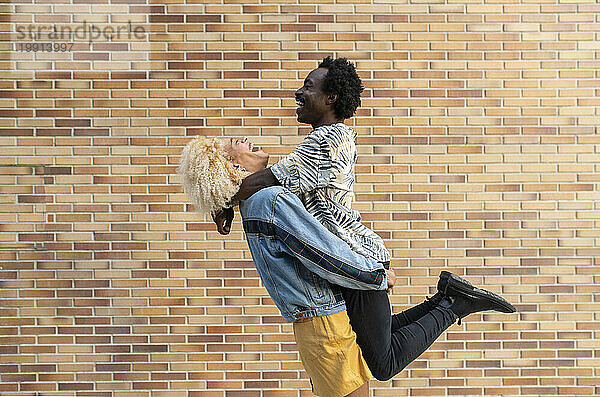 Cheerful transgender woman lifting boyfriend in front of brick wall