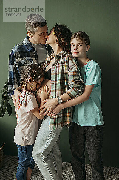 Mann küsst schwangere Frau mit Kindern  die zu Hause vor grüner Wand steht