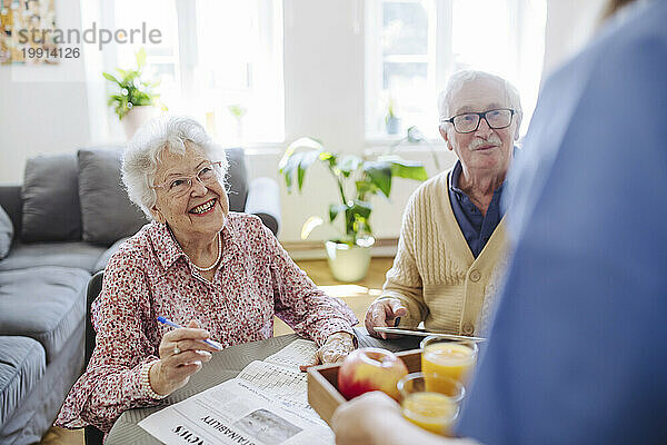 Glückliches älteres Paar mit medizinischem Mitarbeiter  der zu Hause ein Tablett mit Äpfeln und Gläsern Saft hält
