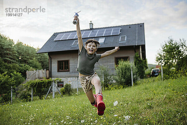 Kleines Mädchen läuft vor ihrem Einfamilienhaus mit Sonnenkollektoren auf dem Dach  in dem ein Windturbinenmodell steckt
